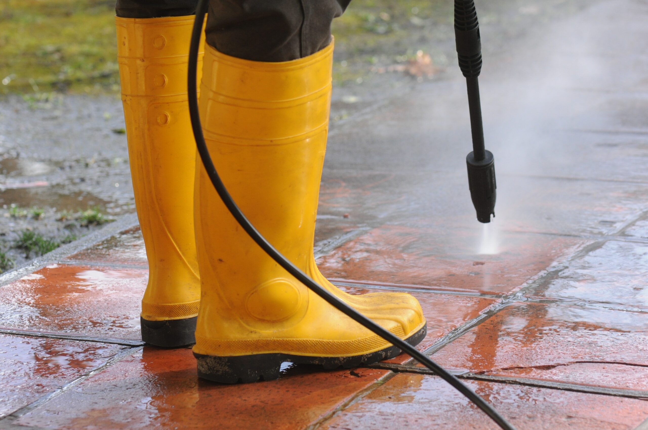 Person wearing yellow rubber boots testing a high-pressure water nozzle on a tiled outdoor flooring. He is one of the many people who wonder, "Can you pressure wash stucco?